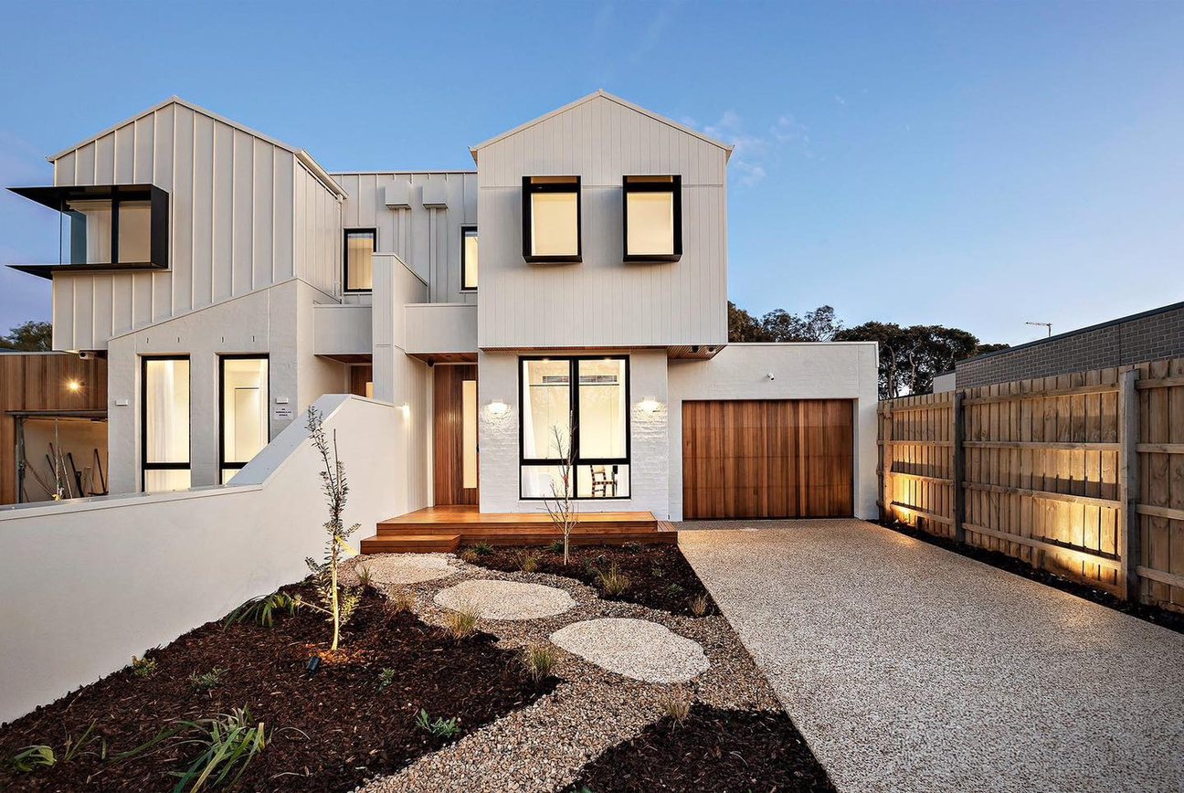 Rosebud duplex at dusk: white and charcoal gabled forms with timber garage doors, gravel driveway and concrete steppers.