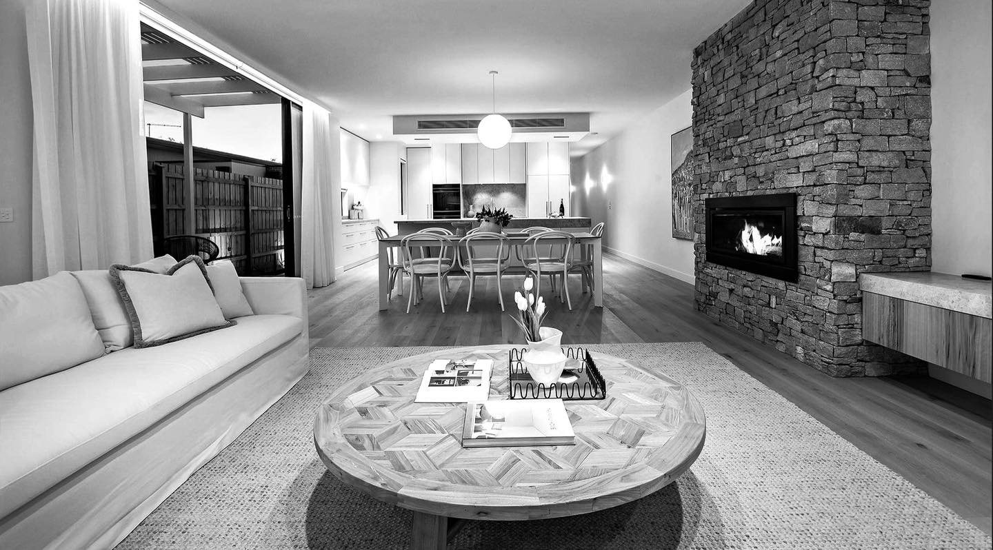 Open-plan living area with a stacked stone fireplace, view through to the kitchen and a marble coffee table on pale floors.