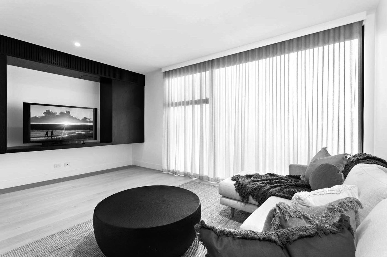 Sitting room with full-height sheer curtains, a linear gas fireplace and a round upholstered ottoman on pale timber floors.